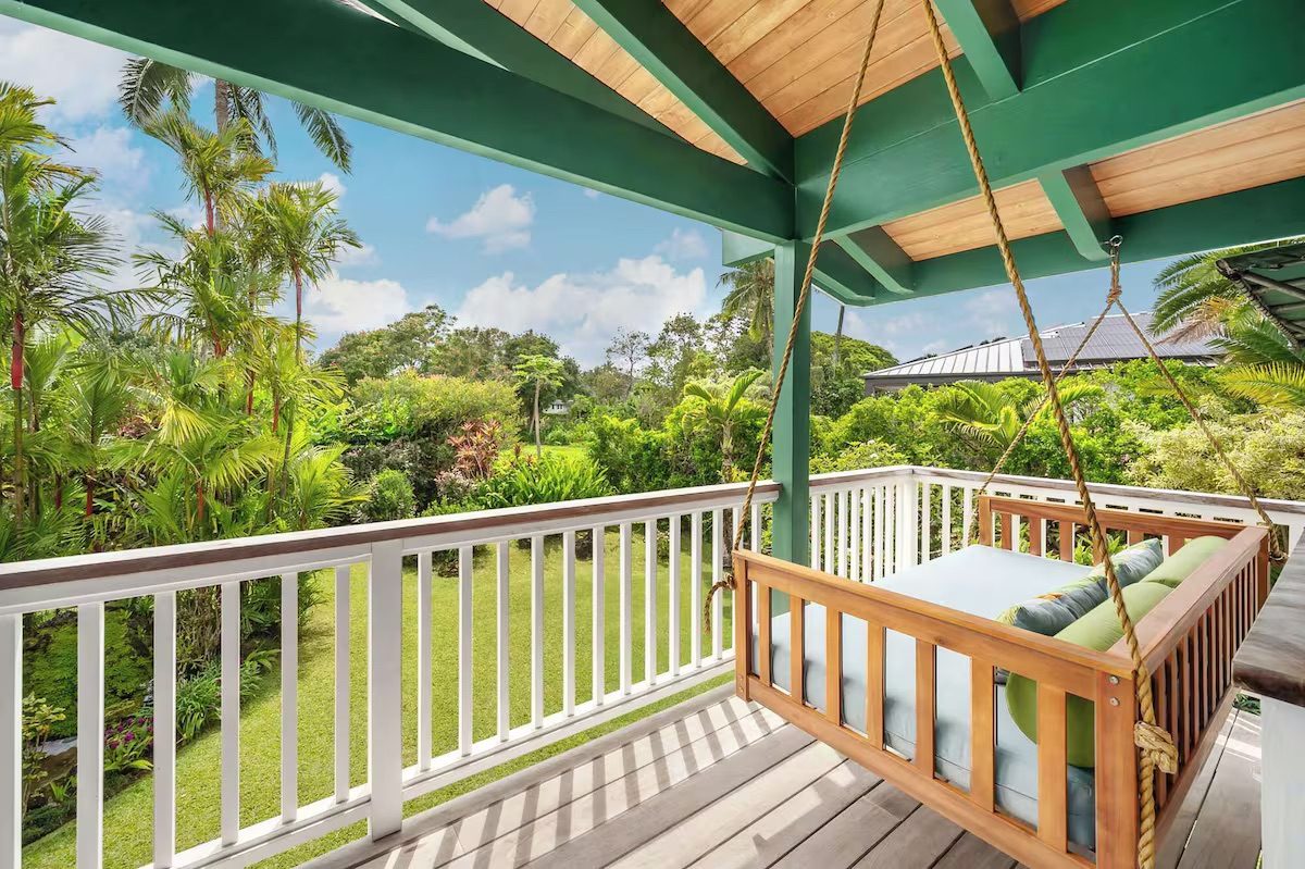 A rope-hung teak swing bed with cushions on the green-and-white lanai, garden and palms beyond.